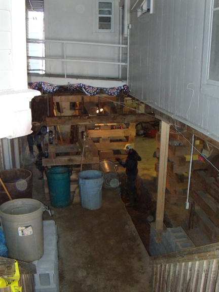 View from the top of the steps between the Shaw and Bird-Werner porches, looking down the walkway between the two buildings. The Shaw kitchen is to the right, where the cribbing is positioned. The trench is for the footing and new block foundation, part of which can be seen just above the lower end of the railing.